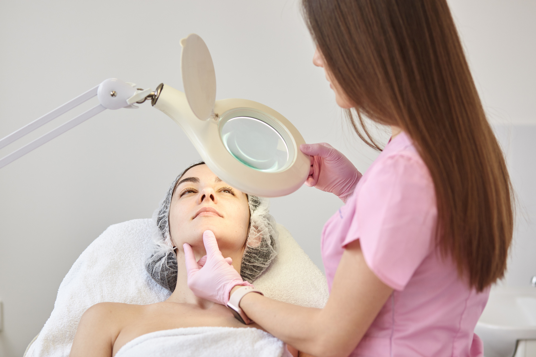 Experienced Young Cosmetologist in Pink Lab Coat Holding Magnifying Glass in One Hand, Switching on Lamp, Touching Womans Face, Assessing Results, Working in Beauty Parlor. Cosmetology Concept.